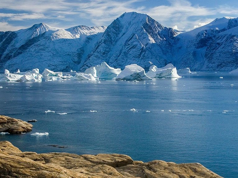 Snowy mountain range with floating icebergs on a calm blue sea, rocky brown shoreline in the foreground