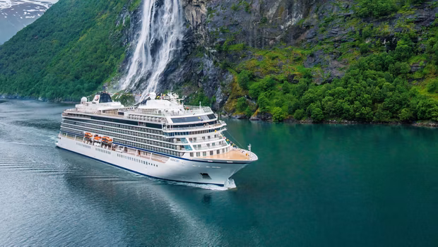 White cruise ship sailing through calm turquoise water near a multi-tiered waterfall and lush green cliffs in a fjord