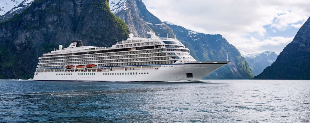 White cruise ship Viking Star sails through a narrow fjord flanked by towering green cliffs and snow-capped mountains.