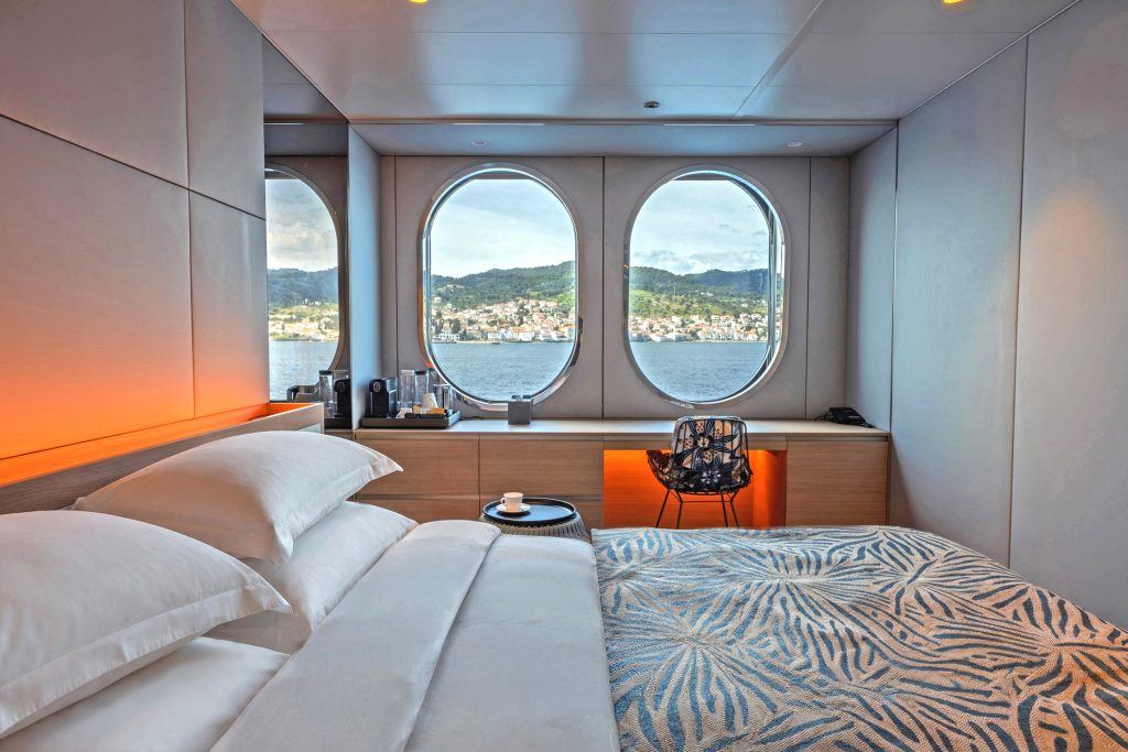 Luxury cabin bedroom with large porthole-style windows showing a coastal town and hills over calm water