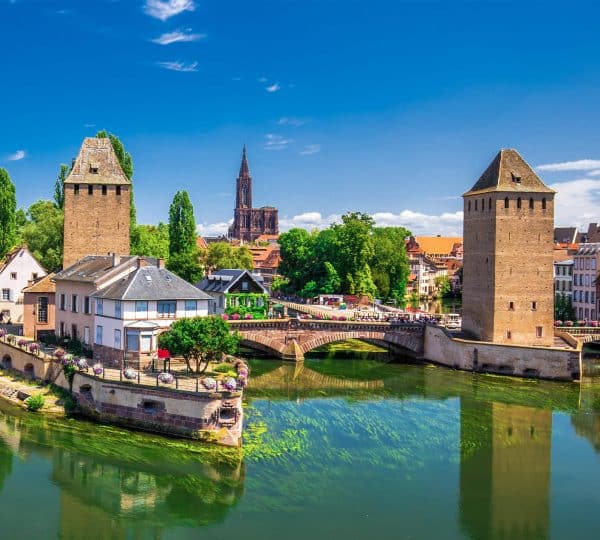 Two medieval towers flank a stone bridge over a calm, green river with colorful town buildings in the background and a church spire visible beyond the trees.