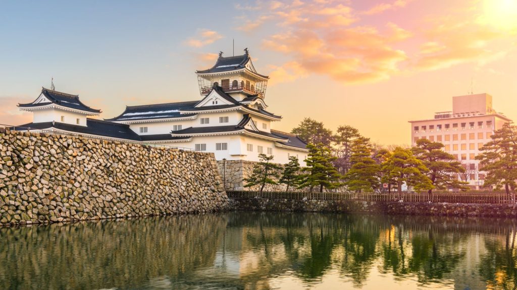 Traditional Japanese castle with white walls and dark tiled roofs beside a stone moat wall at sunset.
