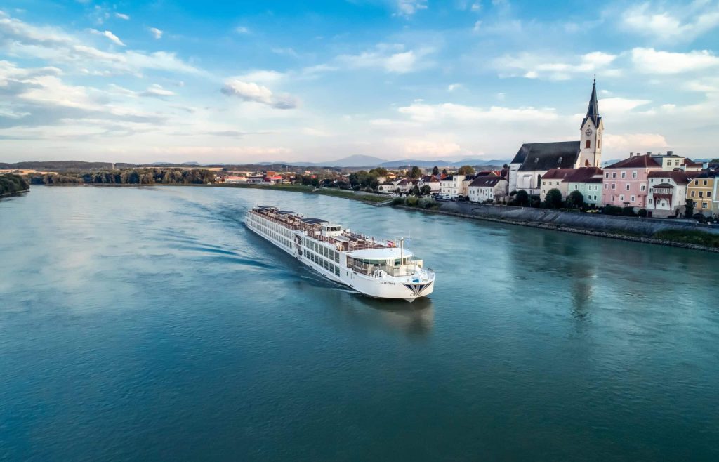 White river cruise boat sails along a calm blue river with a riverside town and a tall church steeple on the right.