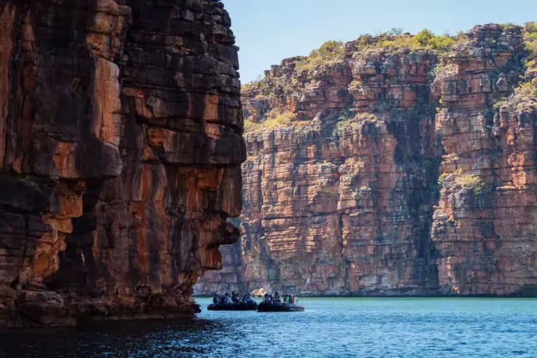 Boats carrying people navigate a calm blue river beside towering red rock cliffs.