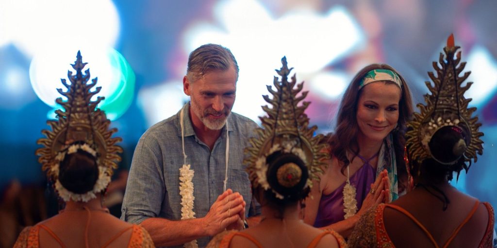 Couple participating in a traditional dance, hands pressed together in prayer amid ornate headdresses and dancers in orange outfits behind them