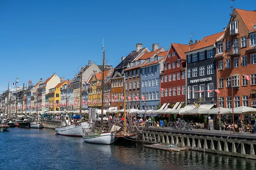 Colorful waterfront row of houses beside a canal with docked boats and people at outdoor cafes on a sunny day (Nyhavn, Copenhagen).