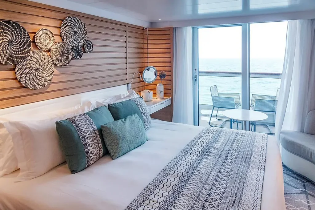 Hotel bedroom with white bedding, teal cushions, and carved circular wall decor; glass doors open to a balcony with ocean view.