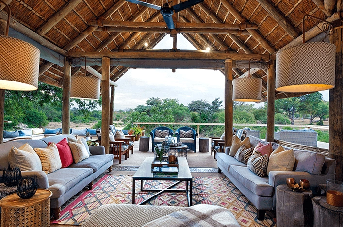 Open-air living room under a thatched roof with gray sofas, colorful cushions, and a central coffee table, overlooking a green landscape.