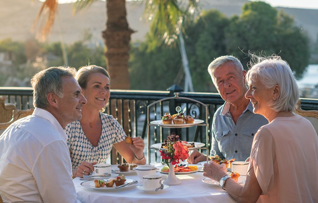 Four adults enjoy tea and light bites at an outdoor terrace with a river view and palm trees in the background.