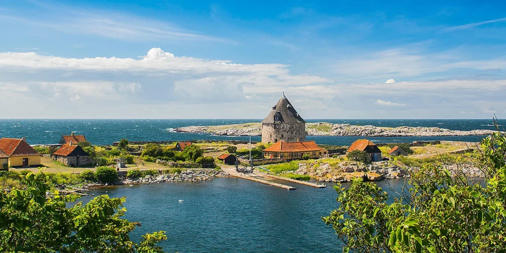 Coastal village with a round stone tower and red-roofed cottages by a blue sea under a bright sky, greenery in foreground.