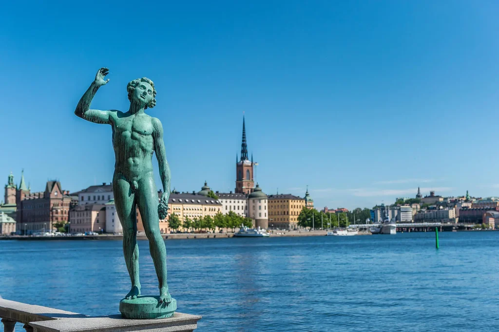 Bronze statue of a nude man raising his right arm, by a river with city buildings in the background.