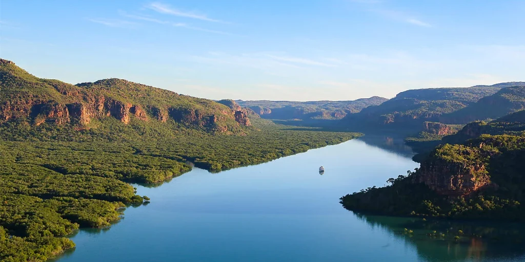 Aerial view of a calm river winding through a lush canyon, with green forested hills and red rock cliffs on both sides; a small boat sits in the middle of the water.