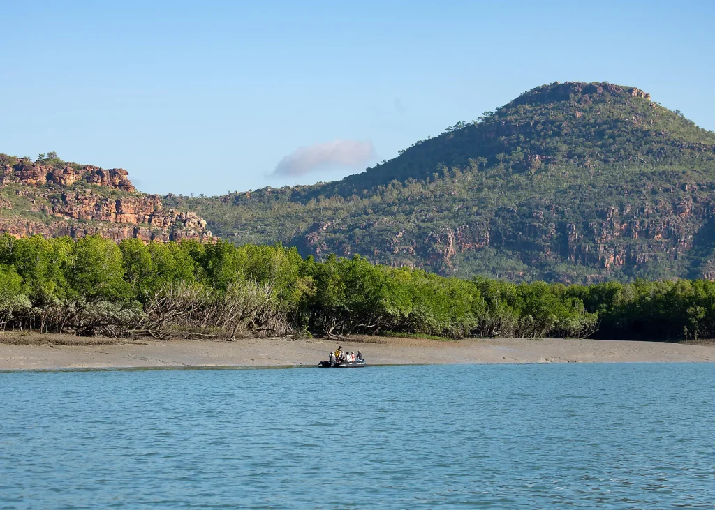 Group of people in an inflatable raft on a calm blue river, with forested hills and a clear sky behind them.