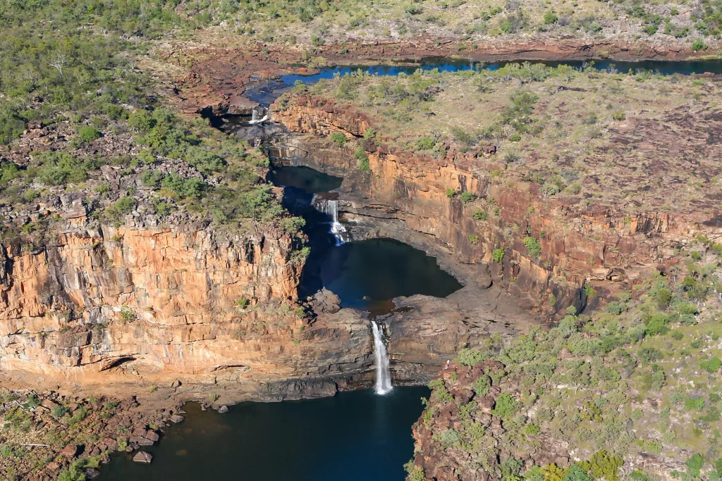 Aerial view of a canyon with layered red rock, a waterfall feeding a dark blue pool below surrounded by sparse vegetation.