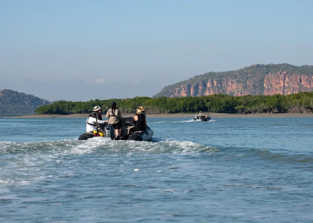 Group of people in life jackets riding an inflatable motorboat on calm blue water with a forested coastline and rocky hills in the distance.