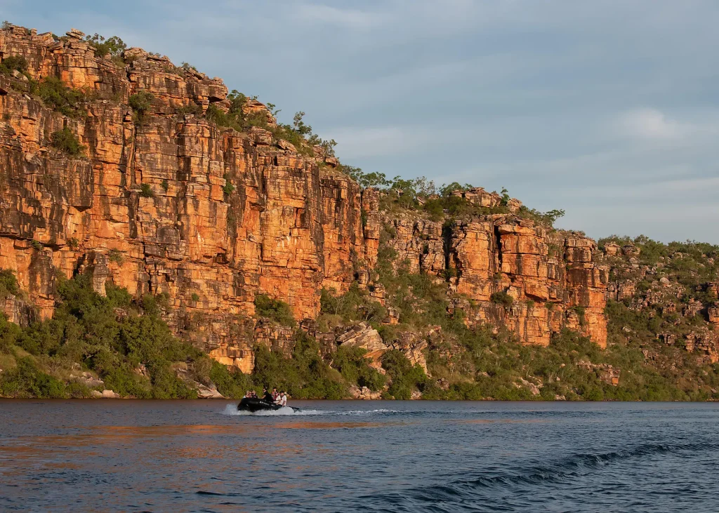 Motorboat with several passengers glides across a calm lake beside a tall, orange-brown sandstone cliff wall with sparse trees.