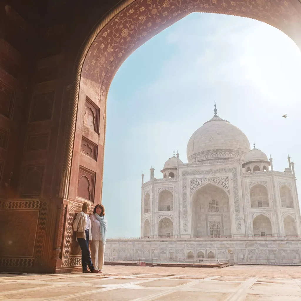 Two women stand under a carved arch framing the Taj Mahal against a bright blue sky.