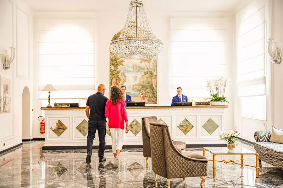 Two guests walk toward the hotel reception desk in a bright, elegant lobby with a crystal chandelier above.