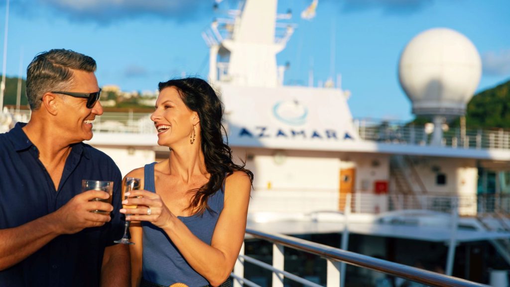 Couple on a cruise ship deck toasting with glasses, smiling at each other with a blue sky and ship behind them