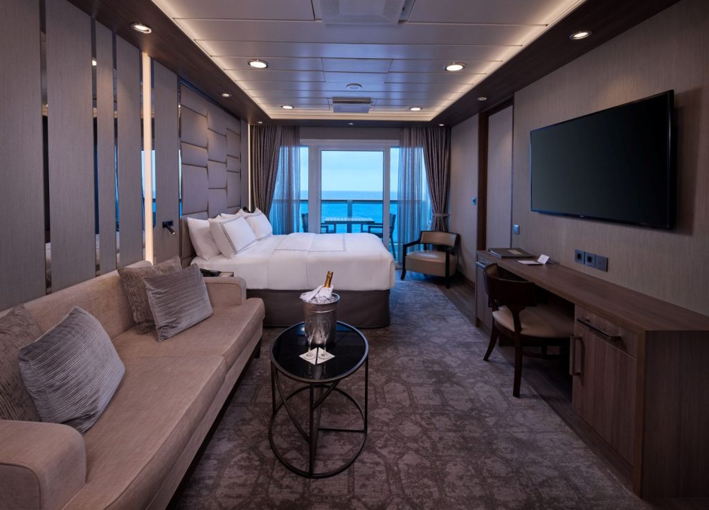 Luxurious hotel room with a king bed, beige sofa, and a balcony overlooking the ocean.