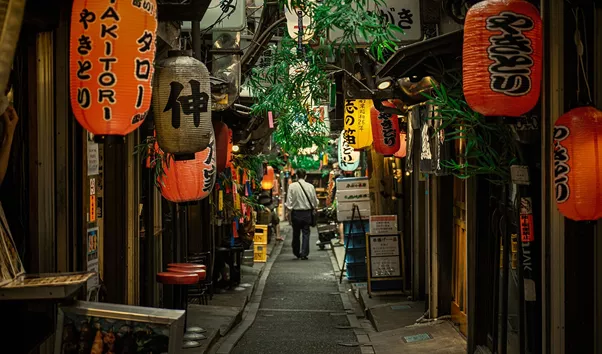 Narrow, lantern-lit alley in Japan with orange paper lanterns overhead, wooden storefronts, and a person walking away in the distance.