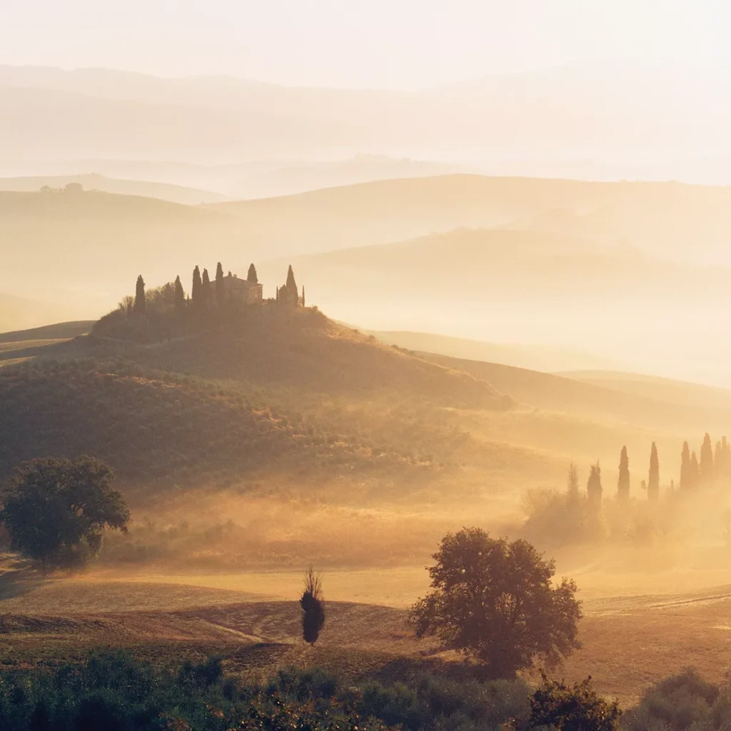 Hilltop house with tall cypress trees on rolling sunlit Tuscan hills at sunrise