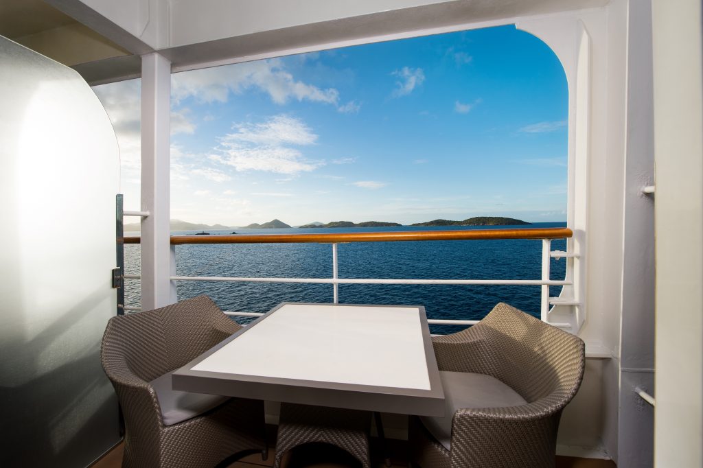 Outdoor seating area on a cruise ship balcony with wicker chairs and a white table, overlooking the blue ocean and distant islands.