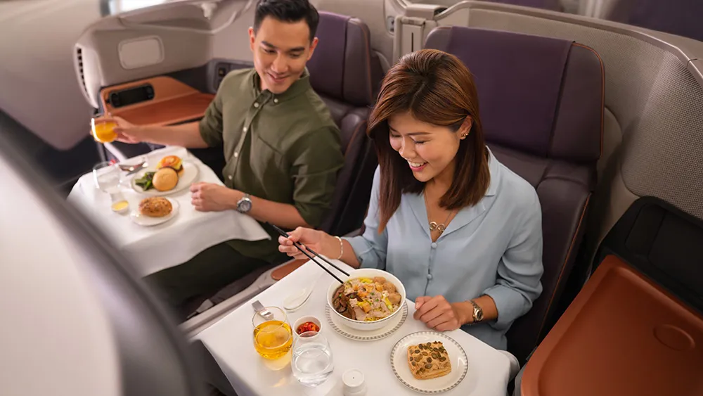 Couple in business class enjoying an in-flight meal, woman using chopsticks to eat a bowl dish while man smiles with orange juice nearby