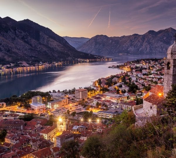Evening view of a coastal town along a calm bay, with lights on streets and red-roofed houses on a hillside, mountains in the background.