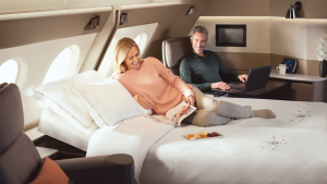 Couple relaxing on a luxury private jet bed: woman reads a magazine while man works on a laptop nearby.
