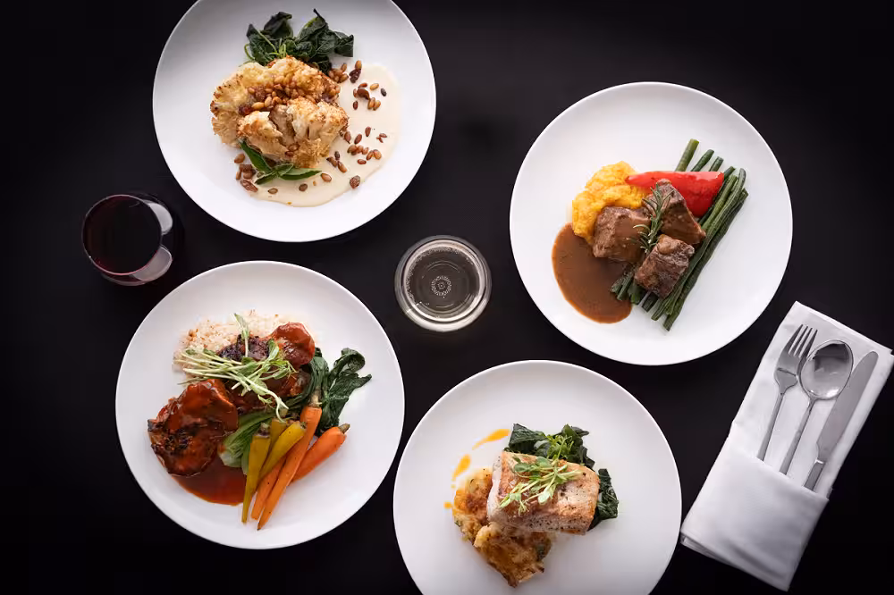 Overhead view of five plated dishes with meats, vegetables, and sauces arranged around a central glass on a dark table.