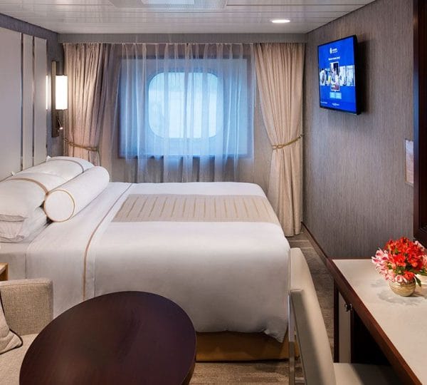 Hotel cabin with a large white bed, sheer curtains, and a wall-mounted TV on the right.