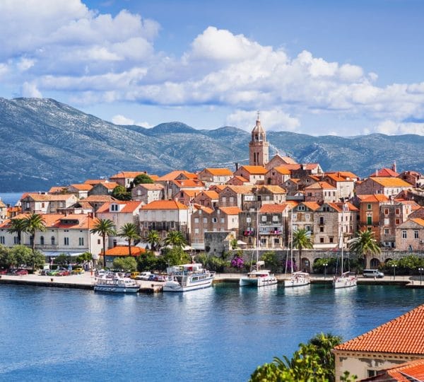 Coastal town with orange-tiled roofs along a calm blue harbor, boats docked in the foreground and mountains in the background, sunny day.