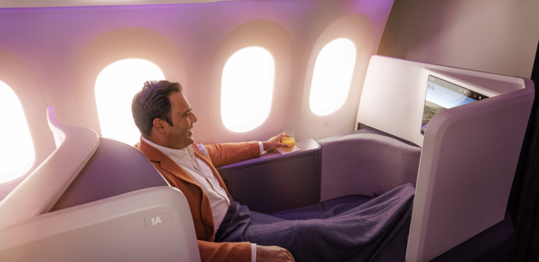 Man reclining in a private airplane cabin seat, holding a glass of orange juice with windows and purple lighting in the background.