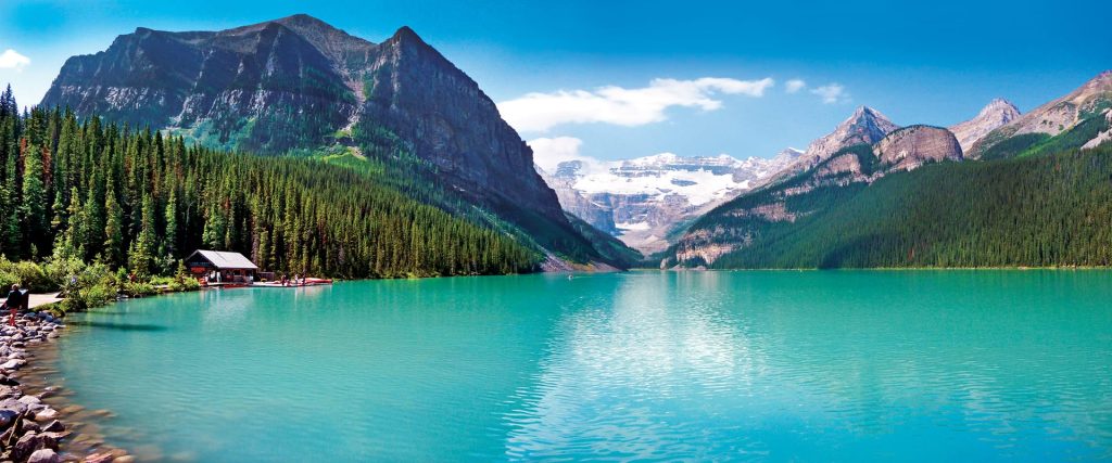 Turquoise alpine lake surrounded by evergreen forest and snow-capped mountains, with a small wooden cabin by the shore.