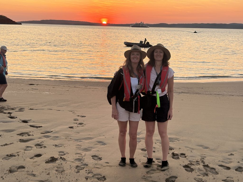 Two women wearing wide-brim hats and red life jackets stand on a sandy beach at sunset, with calm water and a distant cruise ship.